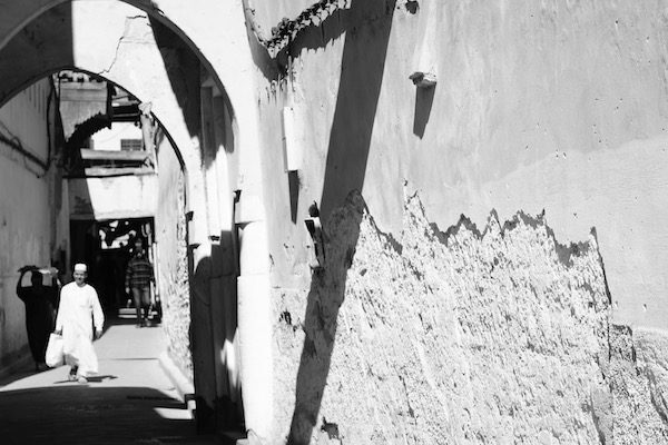 Man in traditional clothing in Fez, Morocco walks down the brightly lit street, midday shadows creasing the ancient pedestrian only passageway. 